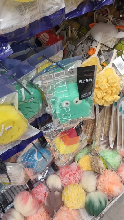 The image depicts a retail display of bath sponges and cleaning supplies, likely in a store in Eilat, Israel, given the context. The scene is densely packed with various items, primarily bath sponges of different colors and textures, including mesh loofahs and foam sponges. Some sponges are individually packaged in clear plastic, featuring labels and branding. Notable items include a green frog-shaped bath sponge with "BATH SPONGE CLEANING SUPPLIES" printed on its packaging and a yellow flower-shaped loofah. In the background, more packaged sponges and what appear to be shower accessories are visible. The lighting suggests an indoor retail environment. No people are visible in the image, and the focus is solely on the product display.