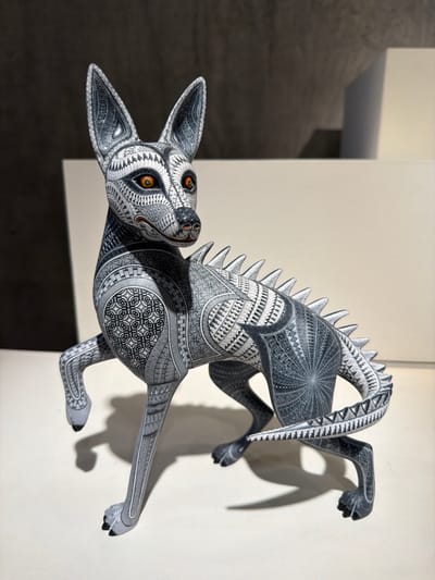 A meticulously carved, monochrome alebrije-style sculpture of a mythical creature is showcased against a neutral backdrop. The figure boasts the body of a canine with intricate geometric patterns etched onto its surface. The creature's back arches with a prominent ridge of spiky scales. Its amber eyes are vividly detailed, contrasting with the overall grayscale palette. The sculpture appears to be leaping or climbing, with its paws outstretched and a curled tail adding to the dynamic pose. The sculpture's shadow is visible on the wall behind it.