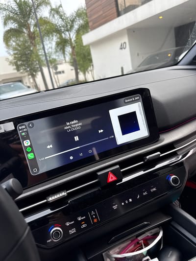 Here is a detailed description of the image:
**Scene:** The image appears to be taken from inside a vehicle, focusing on the dashboard area. The angle is slightly tilted, giving a view of the infotainment system, climate control panel, and some of the exterior environment through the windshield.
**Objects:**
* **Infotainment System:** The central element is a touchscreen displaying a music player interface. It shows a song titled "la radio" by Alleh, Yorghaki from the album "LA CIUDAD" playing on Spotify. There is also an overlay called "Queue & Jam."
* **Climate Control Panel:** To the right of the screen, there's a climate control panel. This includes buttons for A/C sync, temperature control knobs, and controls for front/rear defrost, and driver only setting.
* **Hazard Light Button:** A red triangular hazard light button is visible between the touchscreen and the climate control panel.
* **Dashboard Detailing:** The dashboard shows gray, black and pink accent colors.
* **Other objects:** A UGREEN accessory is visible, along with other personal items.
* **Exterior:** Through the windshield, a building or structure with the number "40" is visible. There are also trees and what seems to be another vehicle.
**Environment:** The time is 6:00.
**Location:** The location in the city of Santa Clara Ocoyucan, Mexico cannot be determined from the image.