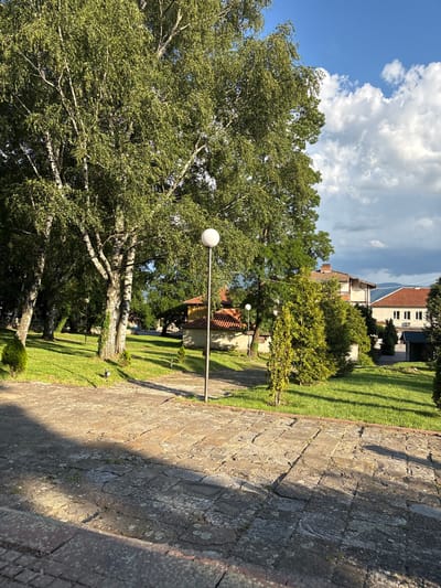 The image shows a park-like setting with trees, grass, and paved walkways. In the foreground, there is a stone pathway with shadows cast by the trees. To the right, a lamppost with a spherical light fixture stands on the grass. In the background, there are several buildings with red tile roofs, and lush green trees surround the area. The sky is blue with scattered white clouds, indicating daytime. The location appears to be a public space, possibly a garden or a courtyard associated with one of the buildings. There are no people visible in the image. The overall impression is one of a well-maintained outdoor area with a mix of natural and architectural elements.