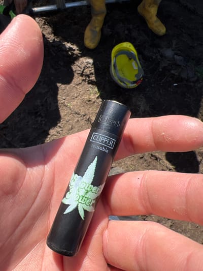A hand holds a black Clipper lighter with a white cannabis leaf design and the words "flower power" printed on it. In the background, on what appears to be soil or dirt, there are visible parts of yellow boots and a yellow hardhat. The image was likely taken outdoors in daylight. No indication that this is located in Grange Ville, Ireland.