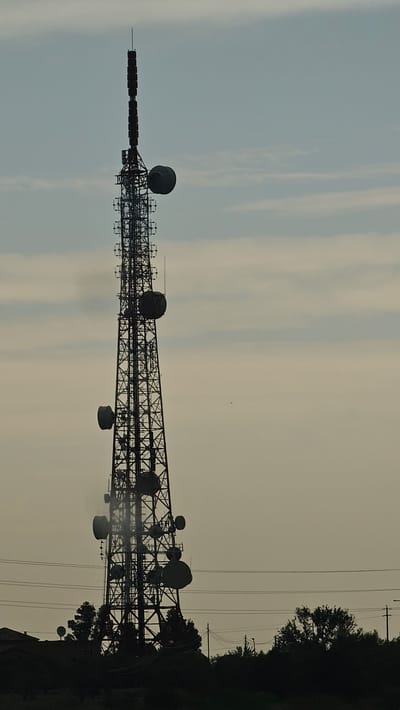 A tall, lattice telecommunications tower stands silhouetted against a hazy, overcast sky. The tower is a complex structure with numerous platforms, antennae, and dish-shaped receivers attached at various levels. At the very top, a narrow spire extends upwards, topped by a single antenna. Multiple rectangular antennae are stacked vertically on the upper section of the tower. Below these, circular or dish-shaped receivers are mounted at different heights on the sides. The tower is framed by dark silhouettes of trees and buildings along the bottom of the image, and power lines are visible crossing the frame horizontally in front of the lower portion of the tower. The lighting suggests either early morning or late afternoon. This is a view of a telecommunications tower located in the municipality of La Muela, Spain.