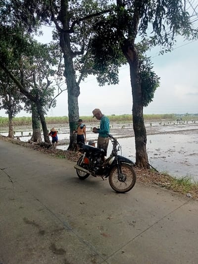 The scene shows a paved road with a black motorcycle parked on the side of the road. A man in a blue shirt and a white hat is standing next to the motorcycle, looking at something on the ground. Two other people are standing in the background, one in a red shirt and one in a blue shirt. All three people are facing a flooded rice paddy field. There is a large tree on the left side of the road and another tree on the right side of the road. The image was taken in Jekulo, Indonesia, at latitude -6.798 and longitude 110.938. The sky is overcast. The image is taken from a low angle. The road is paved and in good condition. The rice paddy field is flooded with water. The motorcycle is a standard black model with a license plate. The people in the image are all wearing casual clothing. The scene is peaceful and quiet.