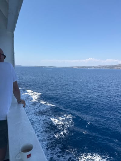 A person stands on the deck of a boat, looking out at the blue sea. The person is wearing a white t-shirt and green shorts and has sunglasses on. A white mug with a red logo is on the railing. The boat is moving, as evidenced by the wake behind it. In the distance, a coastline with hills is visible under a clear blue sky. The location is Nerežišća, Croatia.