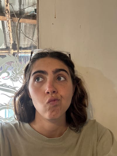 A woman with brown hair and eyes makes a duck face at the camera. She is wearing a beige t-shirt. The background is a white wall with a few marks. To the left is a window with wires and wood, appearing to be under construction. A decorative, possibly glass, panel is partially visible in the lower part of the image. The picture is rotated counter-clockwise.
