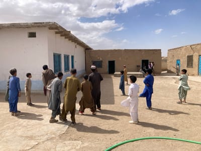 The image shows a group of men and boys standing in the courtyard of a school building in Saranan, Pakistan. The building is constructed of whitewashed mud brick and has blue painted windows.  The men and boys are wearing traditional clothing. The courtyard is dusty and has a green hose lying on the ground. The surrounding area is dry and barren. There are other buildings in the background. The sky is blue and partially cloudy.  The image is taken from a slightly elevated perspective.  The men and boys appear to be interacting with each other, possibly talking or playing. There is a sense of community and activity in the scene.  The image is taken from a slightly elevated perspective.