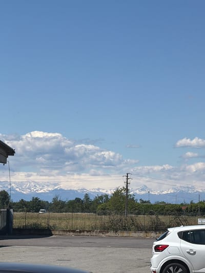 The image shows a view in Gudo Visconti, Italy. The sky is partially cloudy. In the distance, snow-capped mountains are visible. A roadside fence with vegetation separates the foreground from the mountains. Utility poles with power lines are present. Parts of two cars, one white and one dark gray, are visible on the right side of the image. The ground appears to be an asphalt surface, probably a parking lot or road. A sign with Italian text is partially visible near the white car.