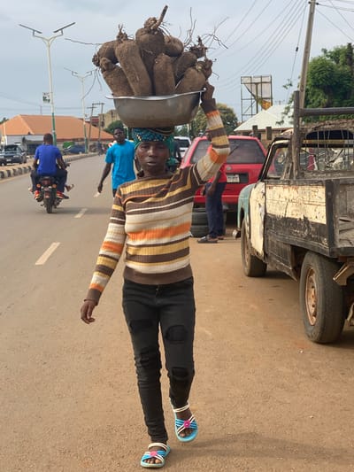 A man carries a large bowl of yams on his head while standing in a street in Kafanchan B, Nigeria. He wears a striped sweater, dark pants, and sandals. The street is paved and has white lines marking the lanes. Other people, including a man on a motorcycle, are visible in the background, as well as vehicles including a truck. There are streetlights and buildings in the background. The sky is overcast.