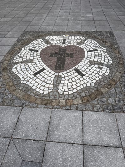 This media file is a top-down, eye-level shot of a decorative mosaic on a sidewalk in Höhenkirchen-Siegertsbrunn, Germany. The mosaic is circular and features a stylized cross shape in the center, made of dark brown and black tiles. This central motif is surrounded by a ring of brown tiles, followed by a larger ring of white tiles arranged in a fan-like pattern. The outer edge of the mosaic is framed by small, grey cobblestones.

The mosaic is set into a sidewalk that transitions from the cobblestone border to larger, square grey paving stones on the right side of the image. The paving stones on the left side of the mosaic are also grey and laid in a grid pattern. The overall impression is of an outdoor public space, likely a pedestrian walkway or plaza.

The lighting suggests it could be daytime, but the overcast sky indicated by the muted colors and the even illumination means it's difficult to determine the exact time of day. There are no people or animals visible in the frame. The focus is entirely on the ground and the intricate mosaic design. The weather appears to be neutral, with no signs of rain or bright sun. No text is visible in the image.