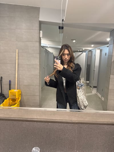 A woman takes a selfie in a public restroom, evidenced by multiple stalls visible in the background mirror. She holds a smartphone in her left hand, her right hand playing with the ends of her hair. She is wearing a black coat, jeans, and a patterned purse hangs over her shoulder. The restroom floor is littered with cleaning supplies, a mop bucket and mop being the most prominent. The wall is tiled in a light grey. There is no contextual information in the image indicating a location in Guazhou County, China.