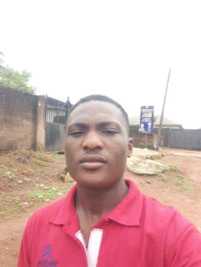 A young Black male is taking a selfie outdoors in Ota, Nigeria. He is wearing a red polo shirt with white trim and a blue logo on the left side of his chest. He is looking directly at the camera with a neutral expression. Behind him, to his left, is a brick wall and a dark metal gate. To his right, a sign with two pictures of men and text in an unknown language is mounted on a pole. The ground is covered in dirt, gravel, and sparse vegetation. The sky is overcast.