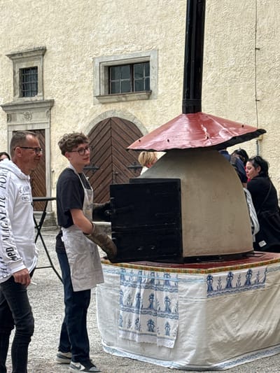 This outdoor scene, set in Častá, Slovakia, depicts people gathered around a traditional outdoor oven, suggesting a baking or cooking event. The main subject is a large, grey, possibly stone or concrete oven with a black metal door. A black chimney pipe extends from the top of the oven, featuring a reddish, triangular metal cap. Draped over the back of the oven and an adjacent surface is a white cloth with distinct blue, folk-patterned designs.
Three individuals are visible. In the mid-ground, a young person with curly brown hair and glasses, wearing a dark t-shirt and a white apron, holds an oven mitt on their right hand, tending to the oven door. Behind the oven, a woman with dark hair, dressed in a dark top, leans over, seemingly assisting with preparations. In the foreground, an older man with short dark hair and glasses wears a white long-sleeved t-shirt with partial text visible, including "MPD MAGNETIC FORCE", "CAMP", and "CHF". He looks towards the activities.
The setting is an old building with a textured, light-colored facade. It features windows with dark frames and grilles, and a large, round-arched wooden door with a radial pattern. The ground is a light, textured surface. The scene is brightly lit, indicating daytime, and conveys a calm, engaged atmosphere around the culinary activity.