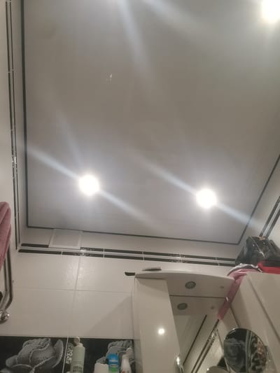 The image captures a high-angle view of what appears to be a bathroom. The ceiling is white and features two recessed light fixtures, both illuminated, creating bright glare. The ceiling has a dark trim around its edges. Below the ceiling, there is a black horizontal band above a tiled surface. The tiles are white and appear to be wet, suggesting a recent shower or cleaning. On the left side of the image, a pink towel hangs from a chrome rack. Further down, there are decorative tiles with a floral pattern. Several bottles of toiletries are visible on a surface in the foreground, including a bottle labeled "URBAN NATURE." On the right side, a stack of items, including a black bag and some folded fabric, are placed on a white surface, possibly a shelf or cabinet. A small round mirror is also visible, reflecting the patterned tiles. The overall impression is that of a well-lit, possibly recently used, bathroom. There are no people visible in the image. The lighting suggests it could be daytime or nighttime, with artificial lights on.