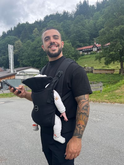 A man is walking outdoors in Ettal, Germany, wearing a black t-shirt and black pants, with a black baby carrier on his chest holding an infant. He is holding a smartphone in his left hand. The background features a lush green hillside covered in trees, with a traditional wooden building with a red roof visible on the slope. The ground is paved with asphalt. The man has a beard and is smiling at the camera. His left arm is visible and covered in tattoos. The infant is wearing white clothing and socks with a red pattern.