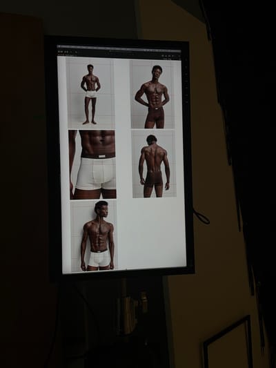 A computer monitor displays a grid of five photographs of a Black male model wearing white underwear. The model is shown from various angles, including full body, torso, and back. The monitor is positioned on a stand, and the surrounding environment is dark, with hints of a yellow wall and a dark curtain or background. The monitor screen exhibits a user interface with menu bars and buttons, suggesting it is actively being used for image editing or review.