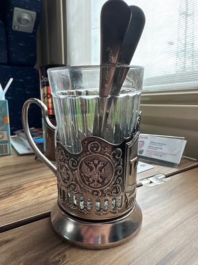 A close-up, rotated image depicts a faceted glass cup held in a silver-colored, intricately decorated metal holder with a handle. Two black plastic spoons are inserted into the glass. The holder features ornate scrollwork and a prominent emblem, possibly a double-headed eagle. The cup is likely for tea or coffee, typical on Russian trains.
The scene appears to be inside a Russian train carriage. In the background, the window shows light streaming through blinds. A business card and other small objects are on the table beside the cup. Below the cup are a blue patterned train seat, and a packaged food item. This is unlikely to be near the city of Zykovo, as it is unlikely that such an item would be used on a suburban train.