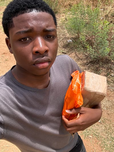 A young man is holding a small cardboard box and an orange plastic bag. He is standing outdoors in a location with dry grass and a small green bush. It is daytime, and the light is bright. The city, Kirana, Nigeria, cannot be confirmed by the image.