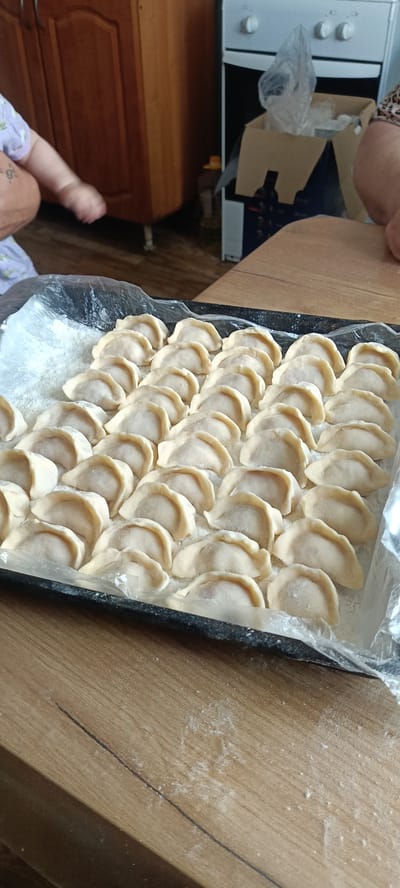In Каменное, Russia, on a wooden table, a black baking tray is filled with numerous uncooked dumplings. The dumplings are arranged in neat rows, dusted with flour. A clear plastic sheet lines the baking tray beneath the dumplings. In the background, part of a kitchen stove is visible, along with wooden cabinets. A cardboard box is placed next to the stove, containing some items. The lower left corner of the image shows a person's arm and leg, suggesting human presence in the immediate vicinity of the prepared food. The scene appears to be set in a domestic kitchen.