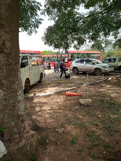The image shows a scene in Ota, Nigeria (latitude: 6.677, longitude: 3.165). Several people are walking towards a row of parked buses. A white minivan is parked close to the bus stop. There is a large tree to the left side of the frame. A large, light gray SUV is parked behind the minivan. There are also red traffic cones on the ground.