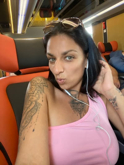 A woman with dark hair and green eyes is taking a selfie on a bus. She is wearing a pink tank top and has several tattoos on her arm and chest, including an eye symbol on her chest. She is also wearing white earbuds. She is sitting in an orange and black bus seat, and other passengers are visible in the background. The bus appears to be traveling through Ninh Hòa, Vietnam, as indicated by the presence of local signage and the style of the bus interior, which is typical of public transportation in the region.
