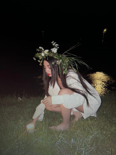 A young woman with long, dark hair and a flower crown is crouching in tall grass near a body of water at night. She is wearing a white dress. Her left hand is reaching down towards a small, glowing object in the grass. The water behind her reflects a distant light. The location is identified as Chotěšov, Czechia.