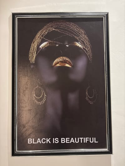 This is a framed piece of art depicting a close-up of a Black person's face against a dark background. The subject has intricate gold braids that frame their face, and their lips are also adorned in gold. The person is wearing gold hoop earrings. The phrase "BLACK IS BEAUTIFUL" is printed vertically in white text on the right side of the artwork. The frame surrounding the artwork has a black and silver ornamental pattern. The artwork is hung on a textured white wall. The location context is Reifild-Ist, Nigeria, but there are no specific visual cues in the image to confirm this location. The lighting appears to be artificial, suggesting an indoor setting. The mood of the artwork is empowering and celebratory of Black identity.