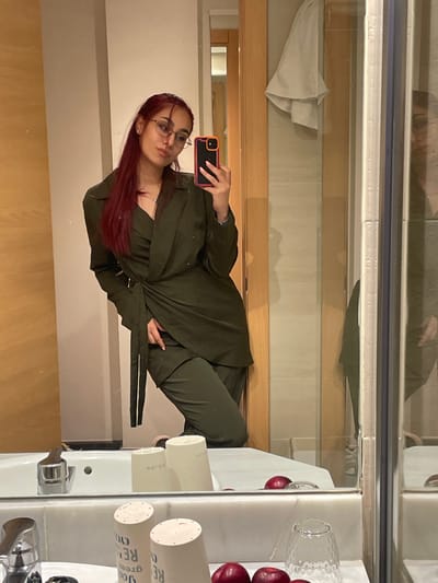 A woman with long, reddish hair and gold-rimmed glasses is depicted taking a mirror selfie in a bathroom, consistent with a hotel setting in Calvià, Spain. She wears a dark olive green, wrap-style blazer over matching trousers. Her right hand holds a black smartphone with an orange and pink-trimmed case, aimed at her reflection. The foreground displays a white marble vanity counter equipped with a chrome faucet. On the counter are two white cylindrical cups, three reddish-purple apples, and a clear glass. The mirror's reflection reveals light-colored tiled walls, a wooden door with a light-colored towel hanging on it, and a glass shower enclosure panel on the right side of the frame.