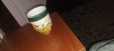 A cylindrical mug with a white interior and a green band near the rim sits on a polished wooden surface. The exterior of the mug is decorated with an image of a woman with red hair in a green dress reclining, with a teacup and swirling lines above her. Small pink heart-shaped details are scattered across the mug's surface. The wooden surface appears to be a table or desk, with a darker, textured carpet visible in the background. The lighting suggests an indoor setting, possibly a room in Zhytomyr, Ukraine, though no specific urban context is discernible from the image itself.
