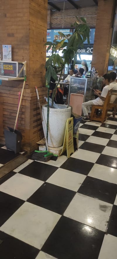 The image shows a black and white checkered floor in a restaurant located at Jalan Gajah Mada 121, Mojokerto, East Java, Indonesia. The restaurant has brick walls, a large plant in a pot, and a black and white checkered floor. There are two mops, a broom, and a trash can. A yellow caution sign stands on the floor next to the plant. A young man is seated at a table to the right. There is a glass case with items behind the man. The restaurant also appears to have a menu on the wall with a picture of chicken.