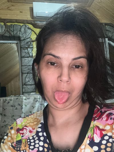 A woman is sticking her tongue out at the camera in a selfie. She has dark hair and is wearing a colorful, patterned shirt. The background appears to be indoors, with wooden paneling on the ceiling and a stone-like wall decoration. An air conditioning unit is visible on the wall. The location is not specified as Kalininskaya, Russia in the image.