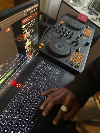 A medium-shot captures a person operating a laptop computer with a DJ controller nearby. The laptop screen reflects the environment, indicating it is in use with a webpage open, possibly a social media platform. The laptop has a backlit keyboard. The person's hand is visible on the keyboard, wearing a ring. The DJ controller is a black and orange model with various knobs, buttons, and two large jog wheels. The scene suggests a setting where music is being created or mixed, and the person is engaged in both computer tasks and DJing. There is no specific information to confirm that the photo was taken in Palaiseau, France.