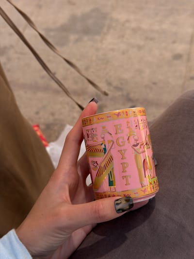 In Giza, Egypt, a close-up shot captures a person's hand holding a pink cylindrical object adorned with Egyptian motifs and hieroglyphs. The object features golden lettering that spells out "EGYPT" vertically. On the object are depictions of figures in traditional Egyptian attire, rendered in gold and colorful accents. The background is blurred, showing fabric textures in shades of brown and gray, suggesting the person is seated or resting. The person's fingernails are painted with a dark, metallic polish. The overall scene is personal and focuses on the decorative object.