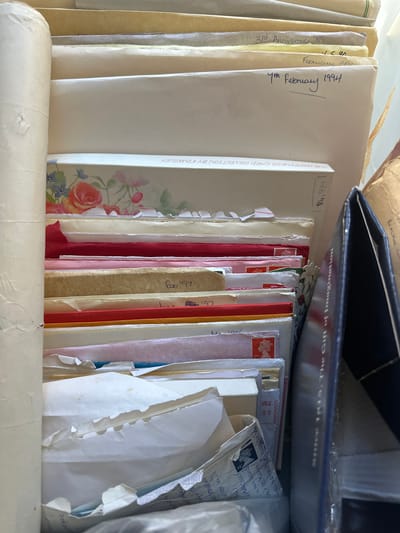 The image displays a close-up, top-down view of a collection of stacked envelopes, cards, and papers. The specific location within Wealden, United Kingdom, cannot be determined from the visual evidence.
The scene features numerous documents, primarily light-colored envelopes, vertically arranged within what appears to be a storage box or compartment. A tightly rolled, light-colored object, possibly paper or fabric, occupies the left margin. Hand-written dates are visible on several envelopes, including "3rd Anniversary '89", "February '90", "4th February 1994", and "Feb '97". One card, visible upside down, reads "THE HAPPINESS CARD COLLECTION BY KINGSLEY". Several items are colored, with prominent red and pink envelopes or cards among the stack. Postage stamps are visible on some envelopes: a red stamp with Queen Elizabeth II's profile marked "1ST", another red stamp, and a blue stamp showing "14P". On the right side, a dark blue or black binder is partially visible, displaying "The Gift of Imagination" on its spine. Behind this binder, a brown paper-wrapped object is partially observable. The papers exhibit varying states of preservation, with some showing creases, tears, and discoloration consistent with age. No individuals are present in the frame.