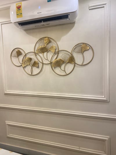 The image, rotated 90 degrees clockwise, presents an indoor scene featuring a white Hisense air conditioner unit and a decorative golden metal wall art piece. The air conditioner, positioned on the left side of the frame, is a split-system indoor unit with the brand name "Hisense" clearly visible on its front. A yellow energy efficiency label is also attached to the lower left portion of the unit.
On the right, a large wall art installation is mounted on a light-colored wall. This piece consists of multiple interconnected golden rings, each containing stylized golden ginkgo leaves. The arrangement of the rings and leaves forms an elegant, organic pattern. The wall itself is adorned with classic white panel molding, creating a structured yet decorative backdrop. The molding appears to outline rectangular sections, enhancing the room's interior design.
No specific activity or event is depicted; the image is a static capture of interior decor and an appliance. The time of day and weather are not discernible from the indoor setting. While the location context is provided as Adenta Municipality, Ghana, the image does not contain specific visual cues to confirm this geographical location.