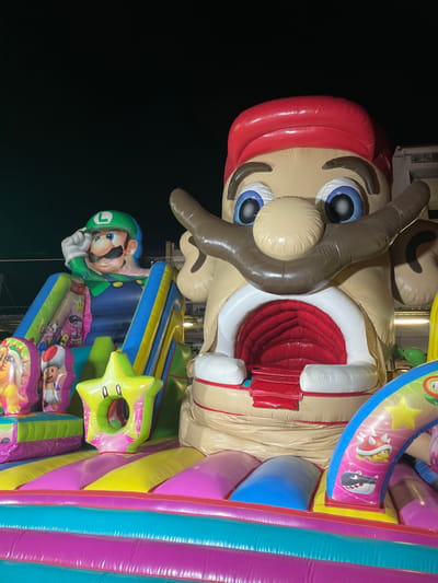 This is a photograph of a large, inflatable, multi-colored bouncy castle themed with characters from the Super Mario video game franchise. In the foreground, the bouncy castle features brightly colored horizontal inflatable sections in pink, yellow, turquoise, and purple. A yellow inflatable star with two black circles for eyes is positioned in the middle ground. To the left, a section of the bouncy castle depicts the character Luigi in his signature green hat and blue overalls, saluting. Behind Luigi, a slide with blue and yellow sides is visible. Dominating the right side of the image is a massive inflatable head of what appears to be a distorted, exaggerated version of a Mario character, with large blue eyes, a prominent brown mustache, and an open mouth revealing red inflatable textures, suggesting a tunnel or entrance. The background is dark, indicating nighttime, with the faint silhouette of a building visible. The image was captured at a public event or attraction in Canet de Mar, Spain.