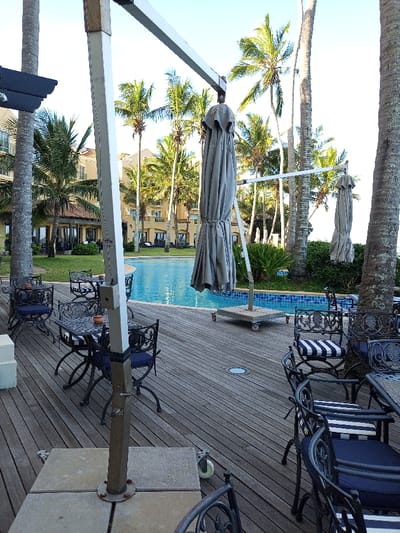 The scene is a resort pool area in Maputo, Mozambique, located at latitude -25.959 and longitude 32.606. The image shows a swimming pool with a large patio area. The patio is furnished with several sets of black wrought-iron chairs and tables, and there are multiple large white umbrellas. Some of the umbrellas are raised and resting on a metal frame with wheels.  The patio is made of wood decking. There are several palm trees surrounding the pool, and there are two large palm trees off to the left, along with a low-rise building in the background. The scene is bright and sunny.  There are no people visible in the scene.