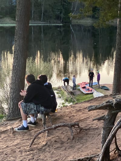 The image depicts an outdoor scene at the sandy bank of a body of water, likely a river or lake, in Vyritsa, Russia. The perspective is from the bank, looking towards the water and the opposite shore.

In the immediate foreground, two individuals are crouched on a reddish-brown sandy slope beneath large pine trees. One person, seen from behind, wears a black hoodie, camouflage shorts, white socks, and dark sneakers with white soles. Their hands are visible, and they appear to be observing the water or something on the bank. Tree roots are prominent on the sandy slope.

Further down the bank, closer to the water's edge, several other people are gathered. An orange and white kayak or canoe is resting on the grass. Among the individuals visible, one is wearing a purple top, another a black top, one a light grey top, and another a blue top, suggesting a group preparing for or concluding a water-based activity.

The water is calm, reflecting the surrounding pine forest and the sky, creating prominent vertical streaks of light and dark. The opposite bank is densely lined with pine trees. The soft lighting, with reflections on the water, suggests either late afternoon or early morning. The scene conveys a relaxed outdoor recreational setting. There is no discernible text in the image.