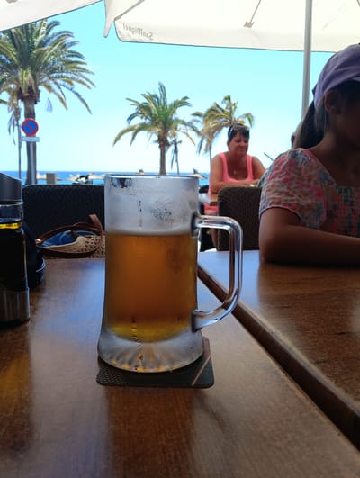 This vibrant image depicts an outdoor dining or bar setting, likely a beachside establishment in Puerto Del Carmen, Spain. The time of day is bright and sunny, suggested by the clear blue sky and well-lit scene.
In the immediate foreground, a large, frosted glass mug filled with golden beer rests on a dark coaster on a wooden table. Condiment bottles in a metal holder are visible to the left of the beer. The table surface is a warm, polished brown.
In the midground, several people are present. To the right, a person, likely a young female, is partially visible from the side, wearing a colorful floral top and a purple baseball cap. Further to the left and slightly behind, a woman with dark hair, wearing a bright pink tank top, is seated at another table, looking towards the right with a soft, content smile. On a table in the mid-left, sunglasses and a straw bag are casually placed.
The background showcases a beautiful coastal landscape. Tall palm trees punctuate the scene against a brilliant, cloudless blue sky. Beyond the trees, the sparkling blue ocean is visible, with distant beach umbrellas dotting the shore. A "no stopping or parking" sign (a red circle with a blue center and a red diagonal line) is discernible near one of the palm trees on the left. Overhead, a large white umbrella provides shade, and the reversed text "San Miguel" is clearly visible on its underside.
The overall scene captures a relaxed, leisurely vacation atmosphere, with people enjoying drinks by the sea.