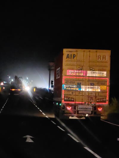 The image shows a nighttime view of a highway in Bohardi Kh, India. The primary subject is the rear of a large, yellow truck with visible advertisements. The truck has license plates and taillights. To the left of the truck, other vehicles are visible on the road, with their headlights illuminating the foggy environment. The road is marked with white lines. The overall scene suggests traffic on a highway at night, with reduced visibility due to fog or mist.