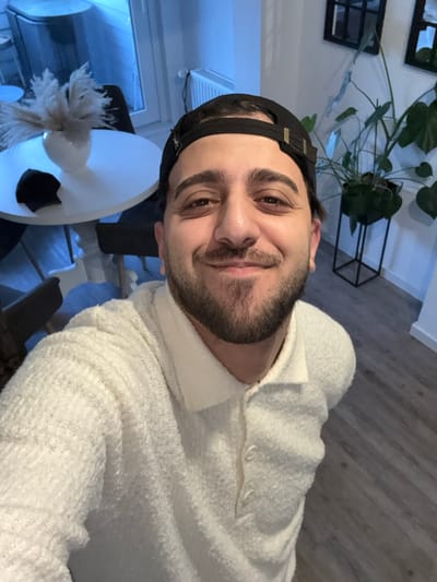 A man with a dark beard and black baseball cap smiles slightly at the camera. He's wearing a white sweater with a textured, nubby appearance. Behind him is a white, round table with a feathery decoration, a black chair, and a window revealing a bluish light. Further back, there's a potted plant on a black stand and a framed artwork on the wall. The background also includes light-colored wooden floors. The lighting seems to be a mix of natural and artificial light.