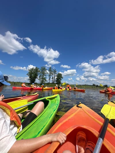 In the Валожынскі сельскі Савет, Belarus, on a bright, sunny day, a group of people are kayaking on a calm body of water. The sky is a vibrant blue, dotted with fluffy white clouds. Trees line the distant shore, with a lush green landscape stretching into the background.
Several kayaks of various bright colors, including red, yellow, and green, are visible. In the foreground, a person wearing a blue cap and an orange life jacket is in a red and yellow kayak. Their arm is resting on the side of the kayak, and their legs are visible inside. Another person in the foreground is in a bright green kayak, wearing an orange life jacket and black compression shorts. Their hand is visible, resting on the edge of the kayak.
Further back, a line of kayaks is proceeding on the water. Individuals in these kayaks are also wearing life jackets. Some are paddling, while others appear to be observing the scenery. The water reflects the blue sky and the surrounding greenery. The overall scene depicts a recreational activity in a natural, picturesque setting.