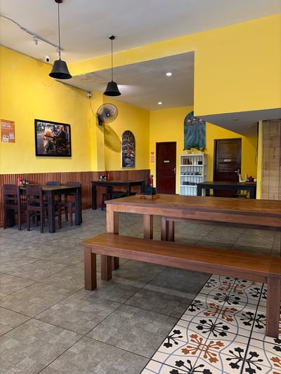 This image depicts the interior of a restaurant with a bright yellow wall and tiled floor. Several wooden tables and benches are arranged in the dining area, suggesting it is open for business. The decor includes framed artwork on the walls, a ceiling fan, and hanging pendant lights. There is also a white shelving unit with items on display.

While no people are actively present in the scene, the setup indicates a place where patrons would dine. The lighting suggests it could be daytime or evening. The style of the restaurant, with its wooden furniture and decorative tiles, hints at a casual and possibly ethnic dining experience.

Visible text includes signage on doors that appears to be in Indonesian, along with a poster on the lower left wall with text that is partially legible, including "FRESH AIR GOOD VIBES". Another sign with red and white markings is near a fire extinguisher. The overall impression is of a welcoming and functional dining space.
