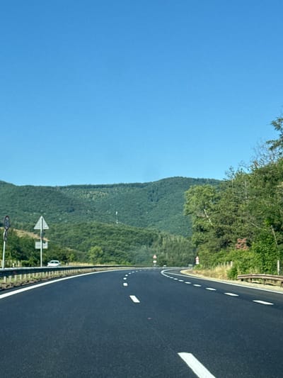 This is a vertical, eye-level, full shot photograph taken on a sunny day. The scene depicts a dark asphalt highway curving to the right, with white dashed lines marking the lanes. The road is flanked by a metal guardrail on the left and a dry, grassy embankment with some trees and bushes on the right. In the distance, rolling green hills covered in dense forest dominate the landscape. The sky is a clear, bright blue. Traffic signs are visible on the left side of the road, including a circular sign with a red border and the number "60" inside, likely indicating a speed limit, and a triangular warning sign. A white car is parked or driving on the shoulder on the left side of the road. A few other vehicles are visible further down the highway. This image appears to have been taken from the perspective of someone traveling on the highway. The location is identified as Dolno Varshilo, Bulgaria.