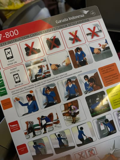 This media file shows a safety instruction card for Garuda Indonesia. The card displays various illustrations depicting safety procedures during a flight, such as how to use an oxygen mask, fasten a seatbelt, and evacuate the aircraft. The illustrations include simplified drawings of people in uniform poses, demonstrating actions like putting on a mask, securing a seatbelt, and navigating an emergency exit.

The card itself is the primary object, with text in both English and Indonesian providing instructions. Visible text includes "Garuda Indonesia," "SAFETY INSTRUCTION CARD/KARTU PETUNJUK KESELAMATAN," "USE OXYGEN MASK FIRST, BEFORE HELPING OTHERS," "KENAKAN MASKER OKSIGEN, SEBELUM MEMBANTU YANG LAINNYA," "KEEP YOUR SEAT BELT FASTEN, AS LONG AS THE FASTEN SEAT BELT SIGN IS ON," "KENAKAN SABUK PENGAMAN, SELAMA LAMPU TANDA KENAKAN SABUK PENGAMAN DINYALAKAN," "FLIGHT MODE ON," "EMERGENCY LANDING," and "EMERGENCY EXIT." There are also icons indicating prohibited actions, such as using electronic devices or not wearing a seatbelt.

The scene is a close-up of the safety card being held, likely on an airplane, given the context of the instructions. The background is slightly blurred, but suggests the interior of an aircraft cabin with muted lighting. The card is held by an unseen person, and the focus is clearly on the safety information presented. The overall impression is one of preparedness and adherence to safety protocols during air travel. The mention of Rawa Burung, Indonesia in the prompt suggests the flight's origin or destination, but this is not directly evident from the image itself.