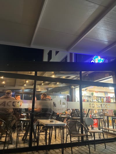 The image captures a nighttime scene of an outdoor seating area belonging to an establishment in Gotse Delchev, Bulgaria. The view is through a large glass storefront or enclosure, revealing approximately six to eight adult males seated at high-top tables with dark metal bar stools. One man in the center-left is observed holding a clear bottle.
The interior of the establishment, visible through the glass, features a bar area displaying numerous bottles on shelves, along with a sign indicating operating hours from "7:00 - 23:00" and "08:00 - 23:00". An ice cream advertisement with a red logo is discernible on the right side of the counter. Warm-toned overhead heating lamps illuminate the space. The glass facade itself bears the white text "MD Shop" on the left and the reversed word "TOBACCO" on the right, suggesting products or services offered.
In the immediate foreground, two empty chairs, one wicker-style and one light-colored metal with a fabric seat, are positioned on a paved ground surface. Above the glass enclosure, a prominent blue neon sign is partially visible, showing characters that resemble "DOMINO'S" from an adjacent or integrated business. The overhead structure of the seating area consists of a light-colored, possibly retractable awning or slatted roof supported by metal beams. In the background, sections of a multi-story building with dark windows are visible against the night sky.