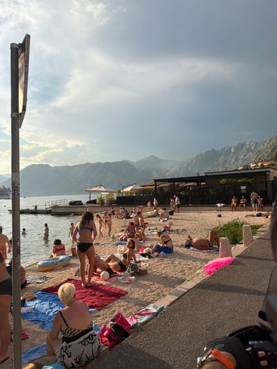 The image depicts a bustling beach scene in Dobrota, Montenegro, on a bright, cloudy day with a mountain range in the background. Numerous people are scattered across the sandy beach and in the shallow water. On the beach, individuals are sunbathing on towels, sitting, and standing. Some are wearing swimwear, including bikinis and swim trunks. Several towels, bags, and a yellow inflatable ring are visible on the sand. In the water, people are wading, and one individual appears to be swimming. Further out in the bay, a large cruise ship is visible. A dark pole with what appears to be a sign attached stands prominently on the left side of the image. The water is calm and reflects the sky. The environment suggests a popular tourist destination during warmer months.