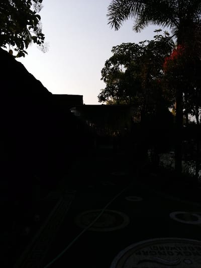 The image depicts an outdoor scene in Kebonsari, Indonesia, at dusk or dawn, indicated by the low light conditions and silhouetted vegetation. The foreground features a dark, paved surface with circular decorative patterns. A green hose lies diagonally across the paved area. In the mid-ground, a low-rise structure, possibly a building or wall, is heavily silhouetted against the sky. The background is dominated by the silhouettes of various trees and palm fronds against a pale, bright sky. The overall impression is one of a garden or courtyard area experiencing very low light.