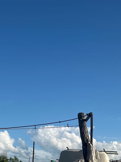 This media file shows a close-up, low-angle shot of a wooden pole against a clear blue sky and some fluffy white clouds. The pole appears weathered and rough, with a diagonal wooden brace attached to it. A dark wire runs down from the pole, with a small green clothespin attached to it. In the background, to the right of the pole, is a white, possibly tarp-covered object supported by a wooden structure, and some greenery is visible at the very bottom right. The scene suggests an outdoor, possibly rural setting in Tobyl, Kazakhstan, on a sunny day with fair weather. The composition focuses on the texture of the wood and the vastness of the sky. There is no visible text in the image.