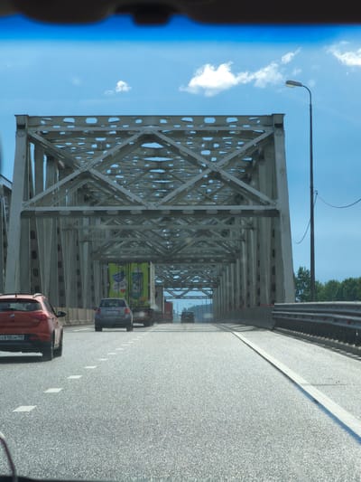This is a photograph taken from the interior of a vehicle, looking forward through the windshield, as it travels across a bridge. The bridge is a large, metal truss structure with a light gray color. The roadway is asphalt, with white lane markings, and a concrete barrier with a metal guardrail on the right side. There are several vehicles on the bridge ahead, including a red hatchback on the left, a gray sedan, and a large green truck. The sky is blue with scattered white clouds. The image appears to be taken in Issad, Russia, a location characterized by its bridges and proximity to water bodies. The scene depicts normal traffic flow across a significant piece of infrastructure.