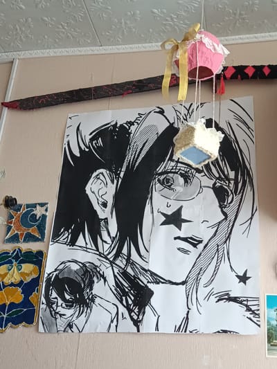 The image shows a wall decorated with a large black and white manga-style drawing of two female faces. Hanging above the drawing is a decorative object made of pink fabric, adorned with lace and a yellow ribbon. Below the pink object, a smaller, cube-shaped decoration with blue and white elements is suspended by strings. To the left of the drawing, there are two small decorative tiles, one featuring a crescent moon and star, and the other with a floral pattern. A prop sword with red accents is mounted horizontally on the wall above the manga drawing. The wall itself is painted a light peach color and has textured white ceiling molding. The floor beneath the wall appears to be a light wood laminate. The scene is indoors, likely in a residential setting. No specific location in the city of Посад, Russia can be determined from the image.